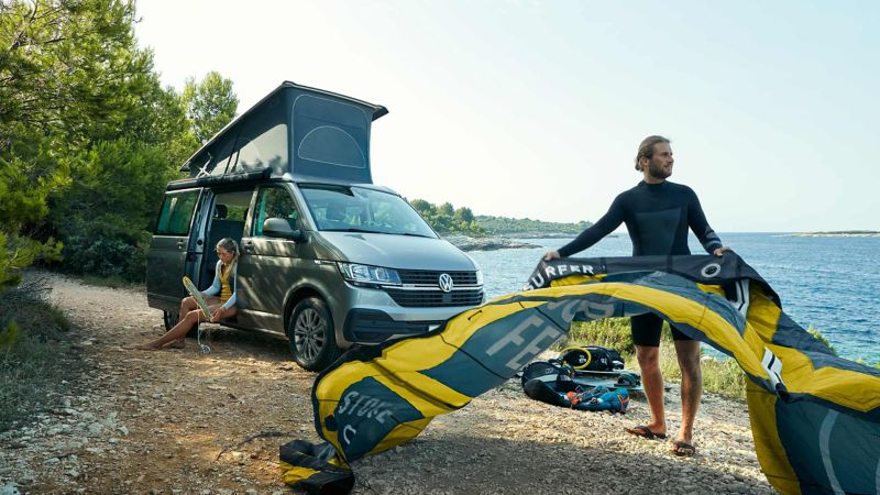 Person flaking kite surf in front of parked Volkswagen California with rooftop tent up.