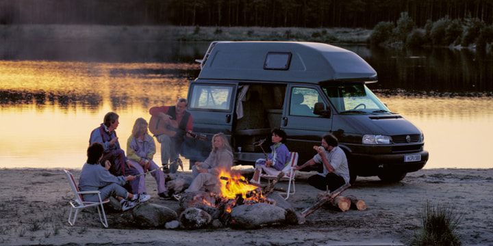 Eine Gruppe Menschen macht abends ein Lagerfeuer vor dem Volkswagen Nutzfahrzeuge California. Das Fahrzeug steht am Strand. Direkt dahinter beginnt das Meer.