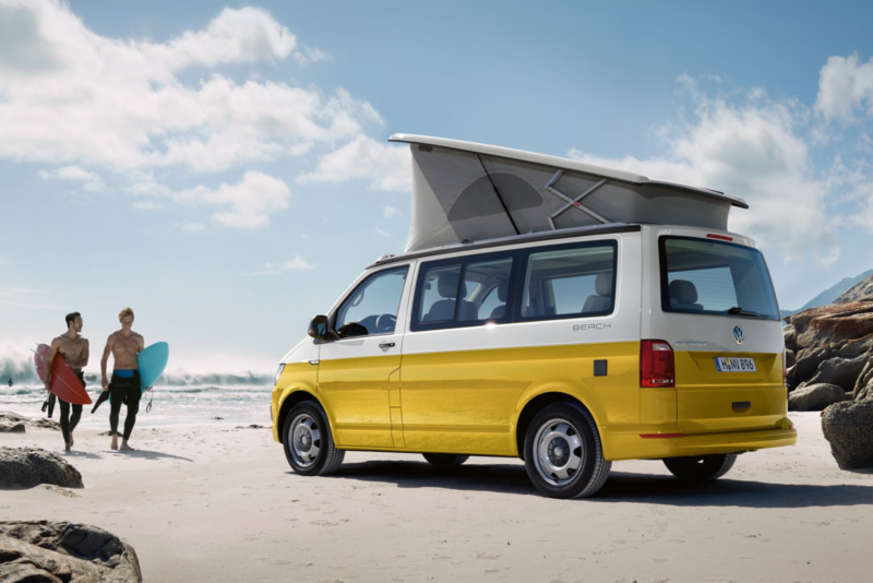 Ein gelber Volkswagen Nutzfahrzeuge California steht bei sonnigem Wetter am Strand. Zwei Surfer laufen darauf zu.