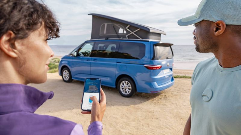 One person holding a mobile phone whilst another looks at a new blue California parked on a beach