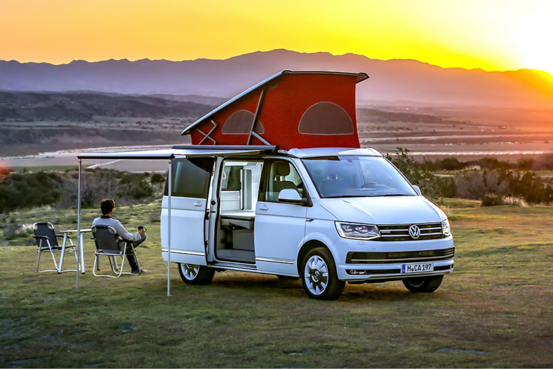 En Volkswagen California med højt tag og udkørt markise holder på en eng. Føreren sidder på en campingstol under markisen og betragter solnedgangen.