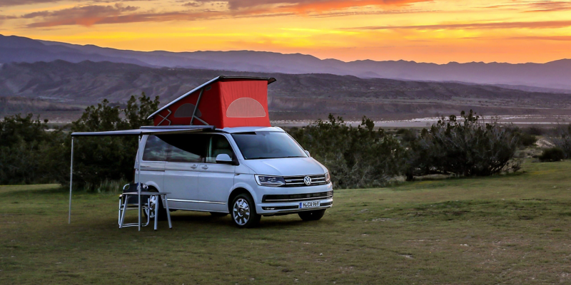 En Volkswagen California med højt tag og udkørt markise holder på en eng. Solen er ved at gå ned.