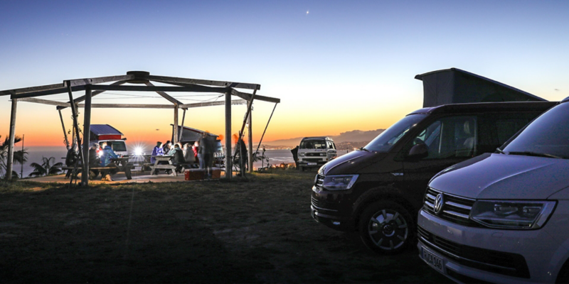 Flere forskellige Volkswagen California-modeller ved en kyst i solnedgangen. I baggrunden kan man ane havet.