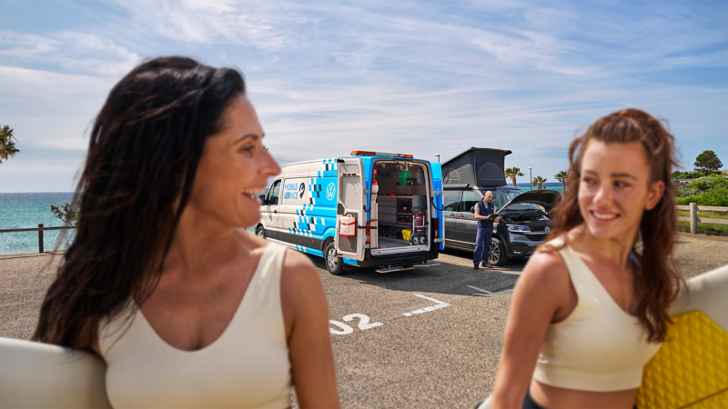 zwei Surferinnen gehen auf einem Parkplatz. Im Hintergrund sieht man einen Mobilen Servicemitarbeiter, der einen VW California Kontrolliert.