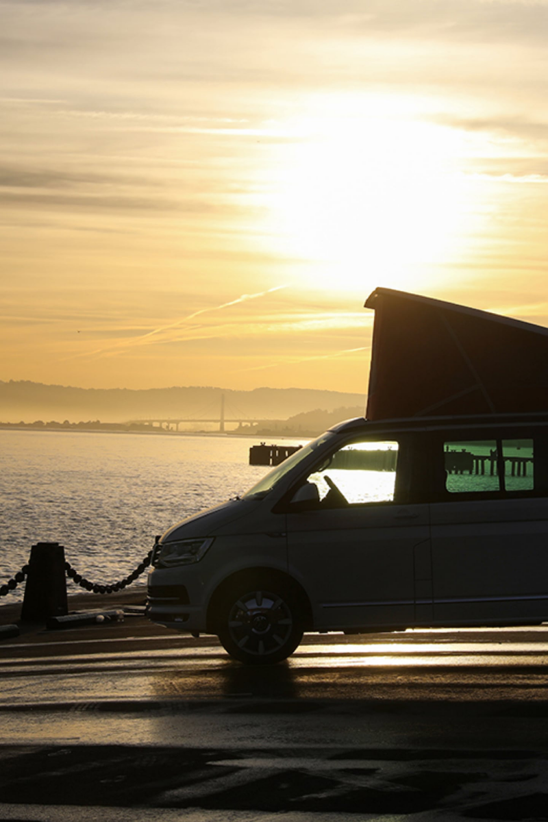 En Volkswagen California holder i modlys på en kyststrækning direkte ved havet i solnedgangen.