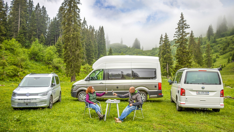 California 6.1, le Grand California et le Caddy California sont garés dans le champ et une femme et un homme se bousculent à la table de camping au milieu.