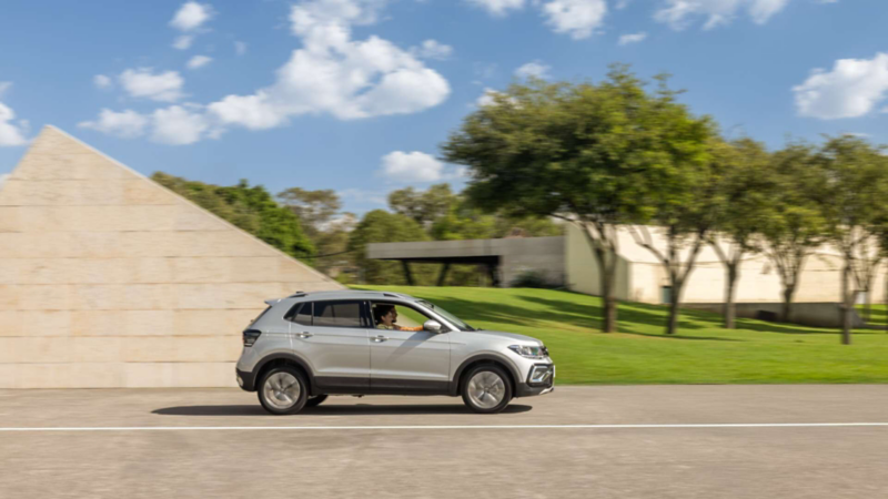 VW Taigun. Vista lateral de SUV de color gris en movimiento sobre carretera junto a edificio moderno.