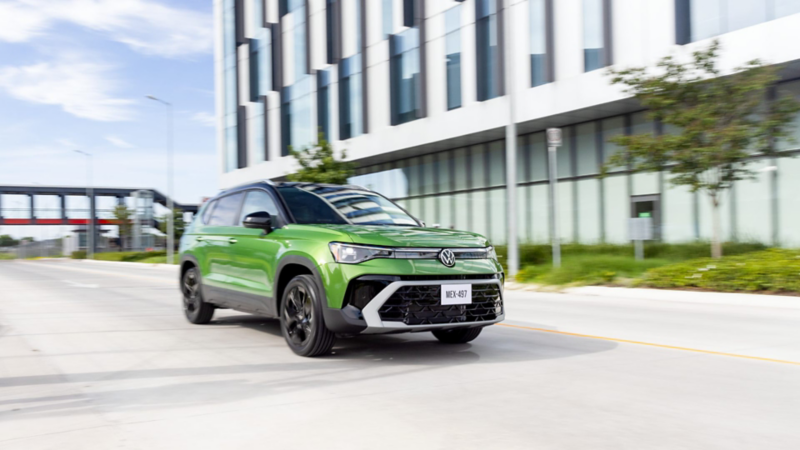Volkswagen Taos color verde circula frente a edificio moderno. SUV compacto con parrilla deportiva y rines negros.