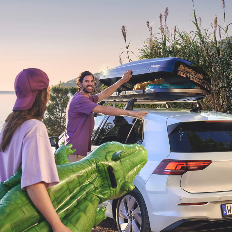 Una chica con un inflable de cocodrilo y un hombre junto a un Volkswagen con un baúl en el techo