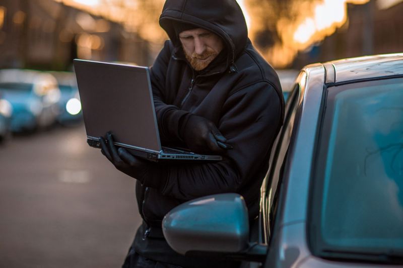 Ein schwarz gekleideter Mann steht mit einem Laptop vor einem Auto und versucht, mit einem manipulierten Funkschlüssel den Wagen aufzumachen.