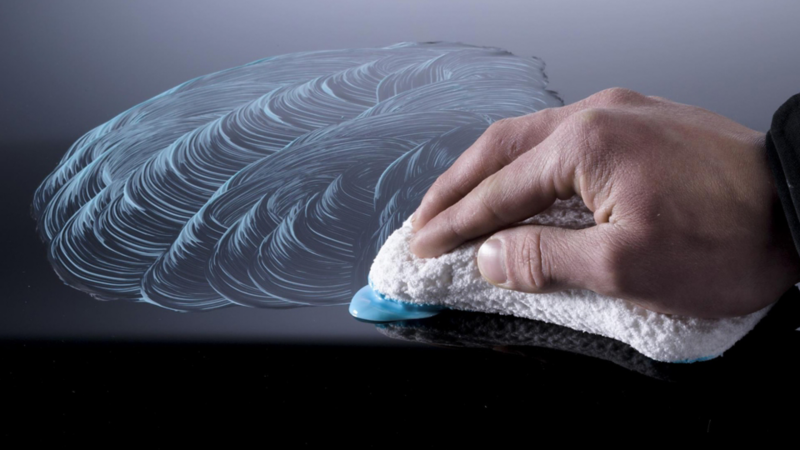 Hand wiping a car surface in circular motions with a microfiber cloth and cleaning product.