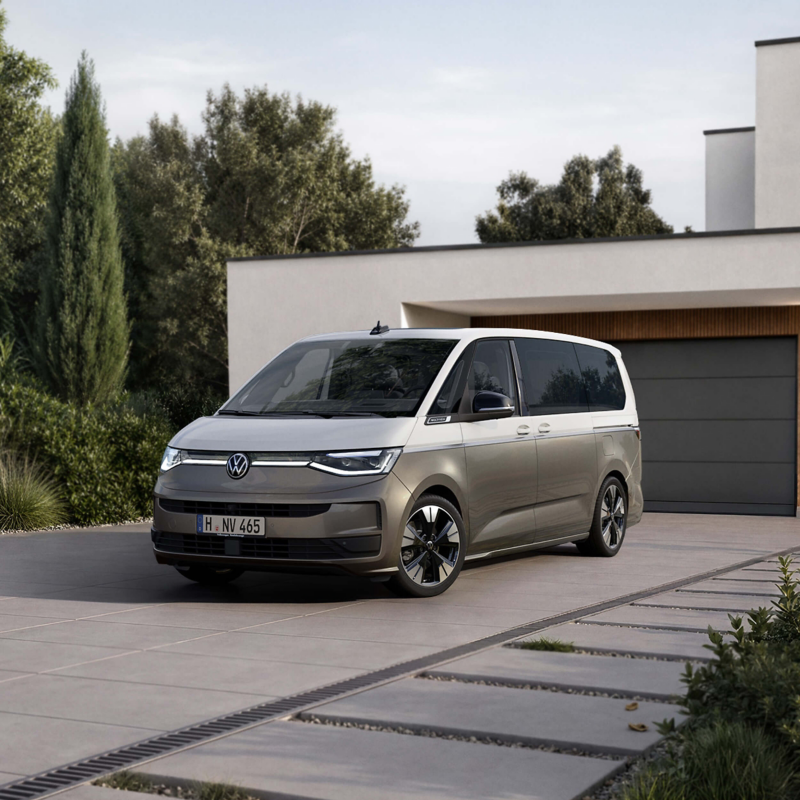 A modern silver van is parked on a sleek driveway in front of a contemporary house, surrounded by lush greenery. The setting is serene and orderly.
