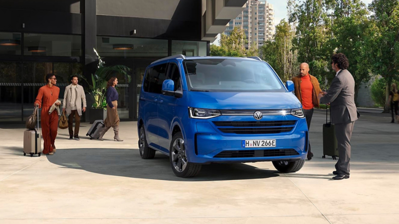 Volkswagen Caravelle bleu devant un bâtiment moderne, des personnes avec des bagages montent et descendent du véhicule.