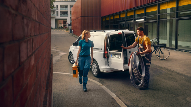 A white Caddy Cargo delivery van is parked outside a building with tradespeople