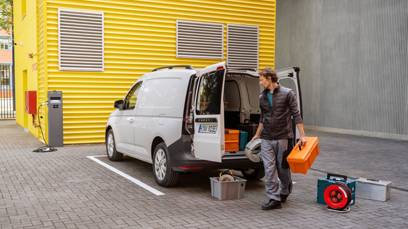 Ein VW Caddy Cargo eHybrid neben an einer Ladestation und ein Mann entlädt Werkzeuge.