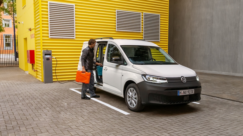 VW Caddy Cargo eHybrid steht an einer Ladesäule neben einer Lagerhalle und wird geladen.