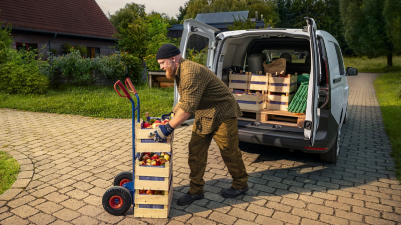 Ein Mann stapelt Apfelkisten aus einer Sackkarre im Laderaum eines Volkswagen Caddy Flexible.