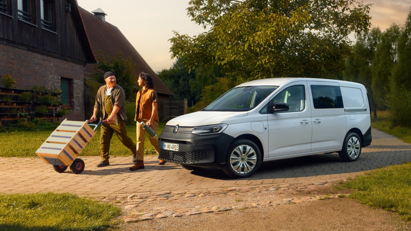 Zwei Personen gehen von einem VW Caddy weg und einer der Personen schiebt eine Sackkarre mit Kisten drauf.