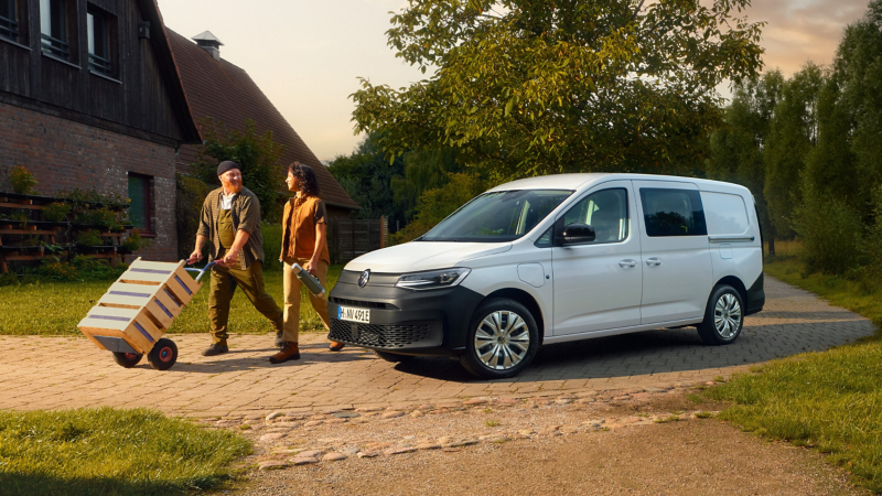 VW Caddy Cargo an einem Bauernhaus. Daneben ein Lieferant mit Obstkisten auf einer Sackkarre.