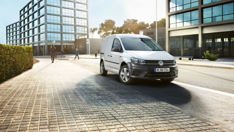  Un utilitaire Volkswagen Caddy blanc est garé sur le pavé d'une rue de ville ensoleillée, avec des bâtiments modernes et des passants en arrière-plan.