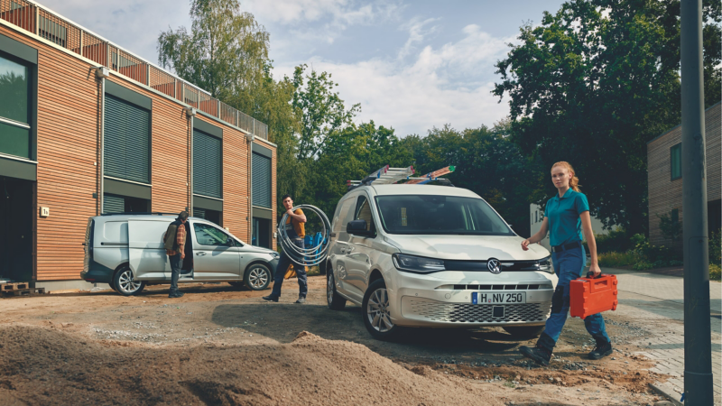 Eine Frau mit Werkzeugkoffer und ein Mann mit aufgerolltem Kabel laufen um ein Volkswagen Nutzfahrzeug auf einer Baustelle.