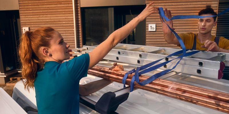 Eine Handwerkerin fixiert eine Leiter auf dem Dach eines VW Caddy Cargo.