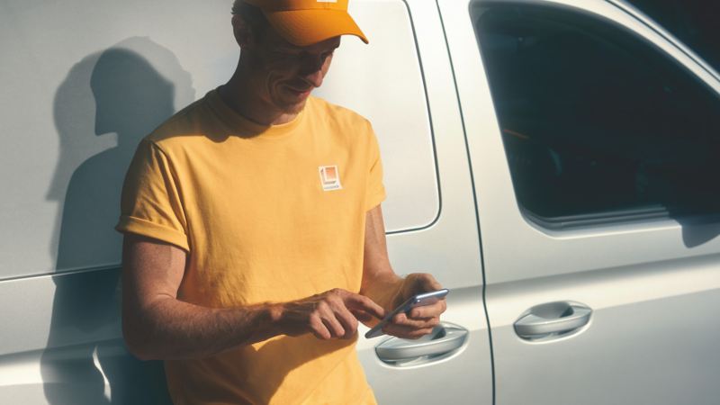 Un uomo con in mano uno smartphone è appoggiato al portellone di Caddy Cargo Volkswagen, visto lateralmente.