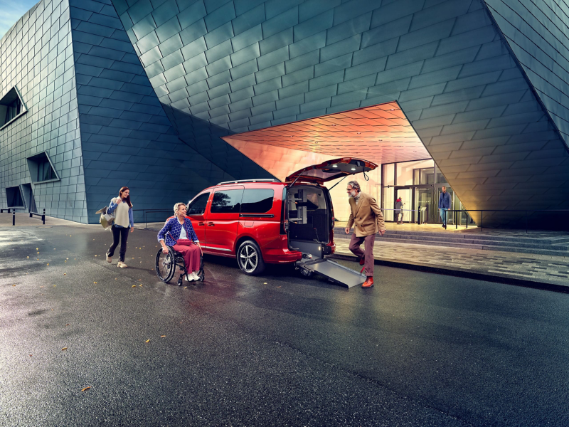 Kobieta na wóżku inwalidzkim podjeżdża do rampy rozłożonej z tyłu czerwonego Volkswagena Caddy.