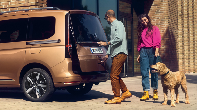 Un uomo chiude il bagagliaio con chiusura servoassistita a comando elettrico di Caddy Volkswagen dopo aver fatto scendere il proprio cane.