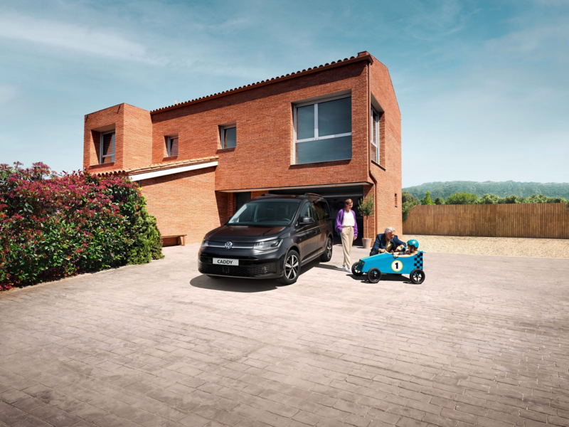 Mężczyzna, kobieta i dziecko w jeździku na podjeździe obok Volkswagena Caddy.
