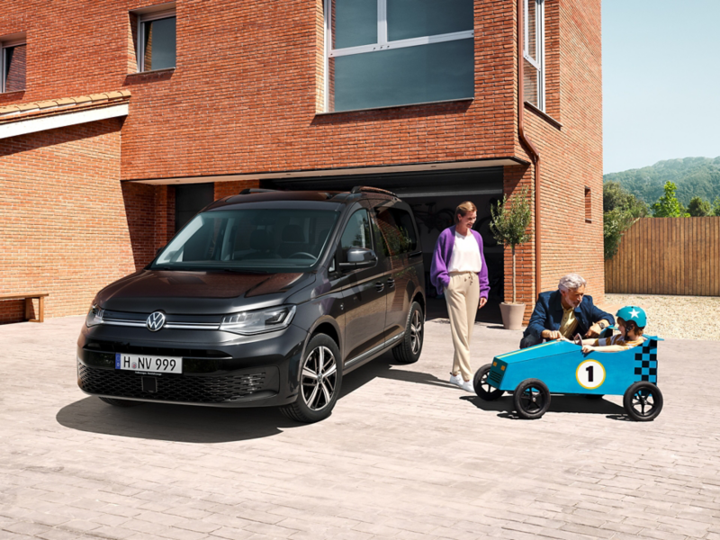 Ein VW Caddy parkt vor einer Garage. Daneben ein Kind in einem Seifenkistenwagen.