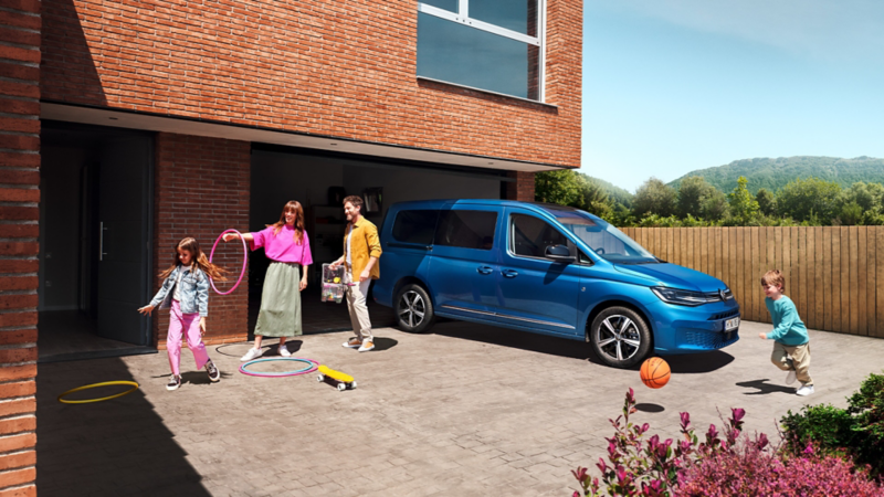 VW Caddy parkt auf einer Auffahrt. Um ihn herum spielt eine Familie.
