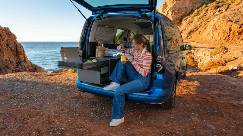 Photo showing a woman sat in the rear section of a VW California pouring a drink against a coastal background. 