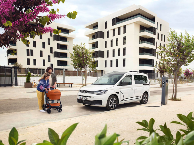 VW Caddy eHybrid steht an einer öffentlichen Ladesäule. Daneben eine Familie mit Kinderwagen.