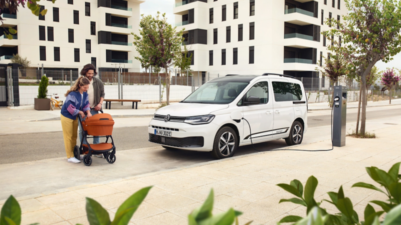 VW Caddy eHybrid lädt an einer öffentlichen Ladestation. Daneben Eltern mit einem Kinderwagen.
