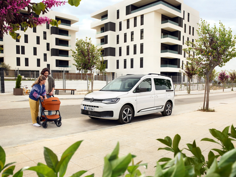 Der VW Caddy parkt in einer Wohngegend und eine junge Familie mit Kinderwagen steht daneben.