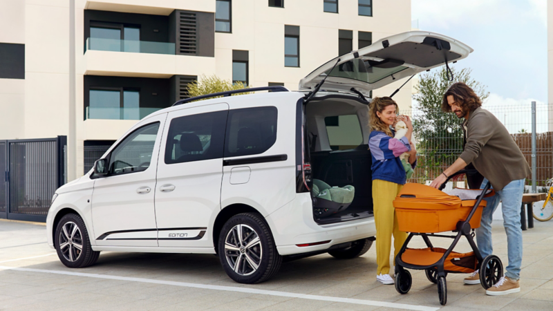 Familie mit Baby und Kinderwagen hinter einem geparkten VW Caddy