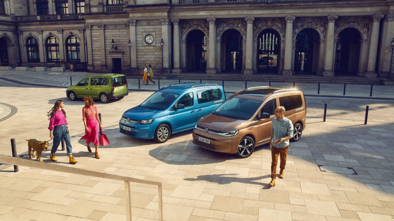 Drei Volkswagen Caddy Range in unterschiedlichen Farben stehen auf einem Platz in einer Innenstadt mit Passanten darum