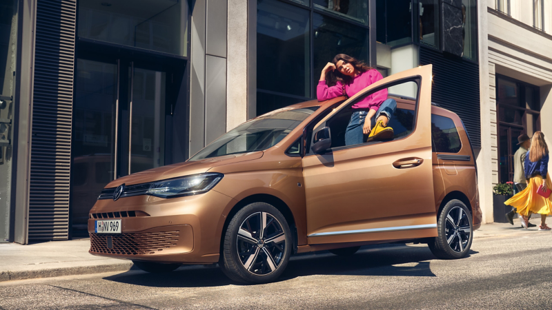Une jeune fille se tient dans l’ouverture de la porte d’une Volkswagen Caddy et a posé le pied sur la fenêtre ouverte.