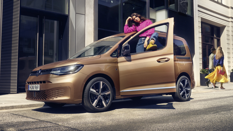 Bronze Volkswagen Caddy parked in city street with woman sitting in open sliding doorway.