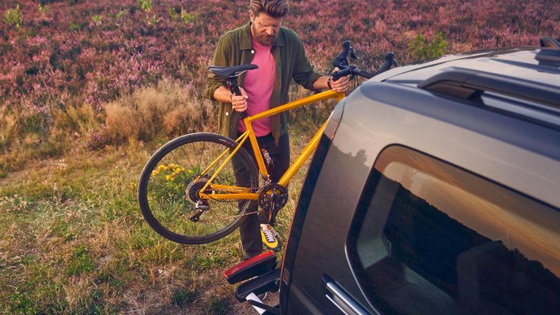 Ein Mann befestigt ein gelbes Rennrad an einem Fahrradträger am Heck eines dunklen Vans in einer ländlichen Umgebung.