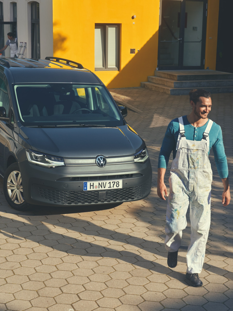 Ein grauer VW Caddy parkt vor einem Gebäude und ein Maler macht sich auf den Weg zur Arbeit