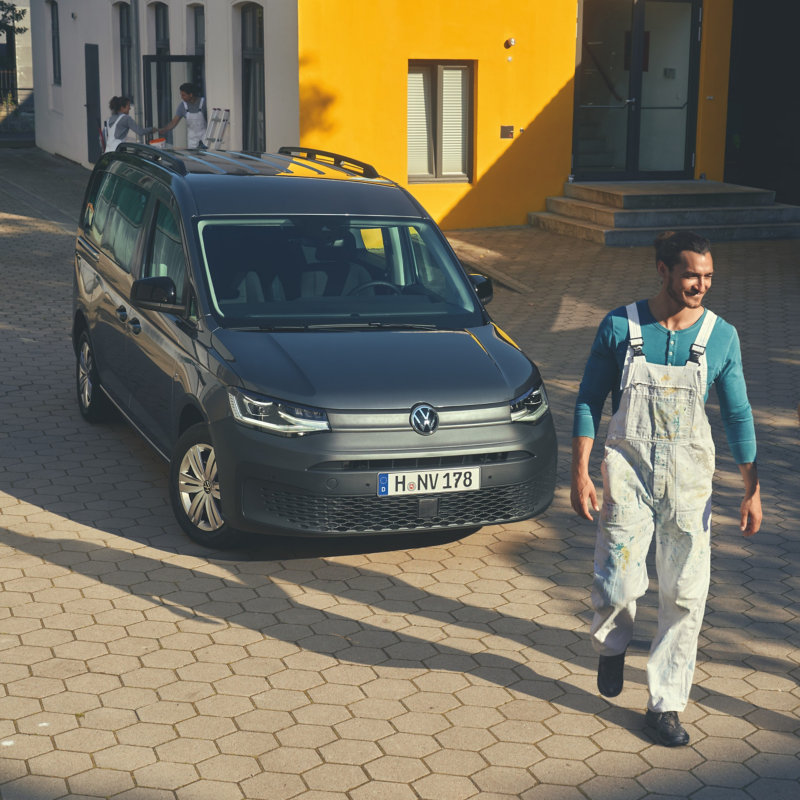 Ein VW Caddy parkt an einem gelb gestrichenen Gebäude. Im Vordergrund ein Maler in Latzhose, der sich vom Fahrzeug entfernt. Im Hintergrund tragen zwei weitere Maler eine Leiter.