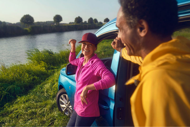 A man and a woman talking in front of a Caddy parked by a river.