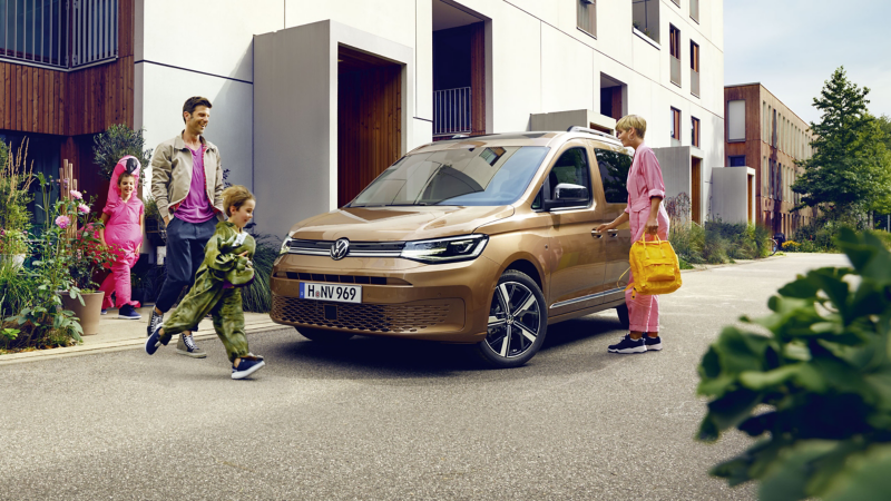 Eine fröhliche Familie mit zwei Kindern in bunten Kostümen läuft an einem sonnigen Tag auf ihren bronzefarbenen VW Caddy zu, der vor modernen Wohnhäusern parkt.