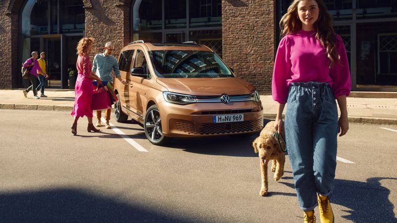 Το νέο Volkswagen Caddy ως οικογενειακό αυτοκίνητο. Ένα ζευγάρι προσεγγίζει το αυτοκίνητο. Πιο μπροστά μια κοπέλα με τον σκύλο της.