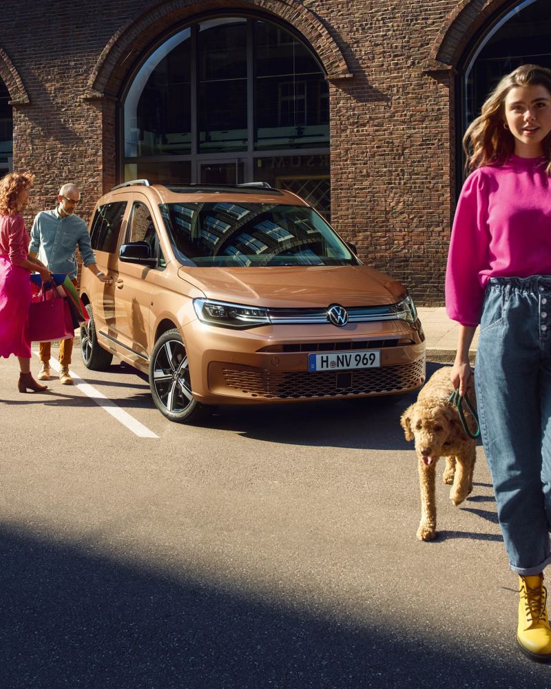 Το νέο Volkswagen Caddy ως οικογενειακό αυτοκίνητο. Ένα ζευγάρι προσεγγίζει το αυτοκίνητο. Πιο μπροστά μια κοπέλα με τον σκύλο της.