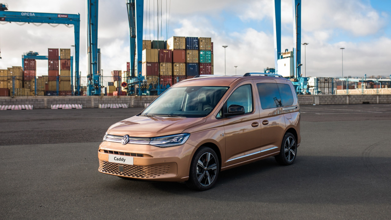 Ein goldgrüner  VW Caddy ist vor einem Hafenterminal geparkt. Im Hintergrund sieht man gestapelte Container und Kräne.