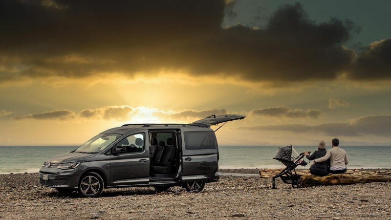 Eine Familie mit Kinderwagen sitzen neben einem VW Caddy auf einem Baumstamm am Meer während die Sonne untergeht