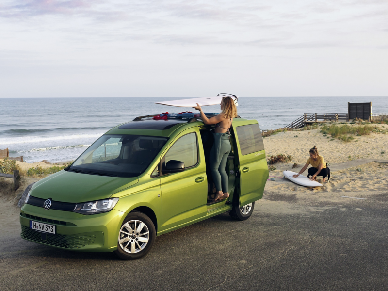 Ein Caddy California steht mit zwei Surferinnen am Strand.